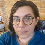 Photograph of a white woman with medium-length, brown hair. The woman is wearing a blue sweater and large-rimmed, tortoiseshell glasses.